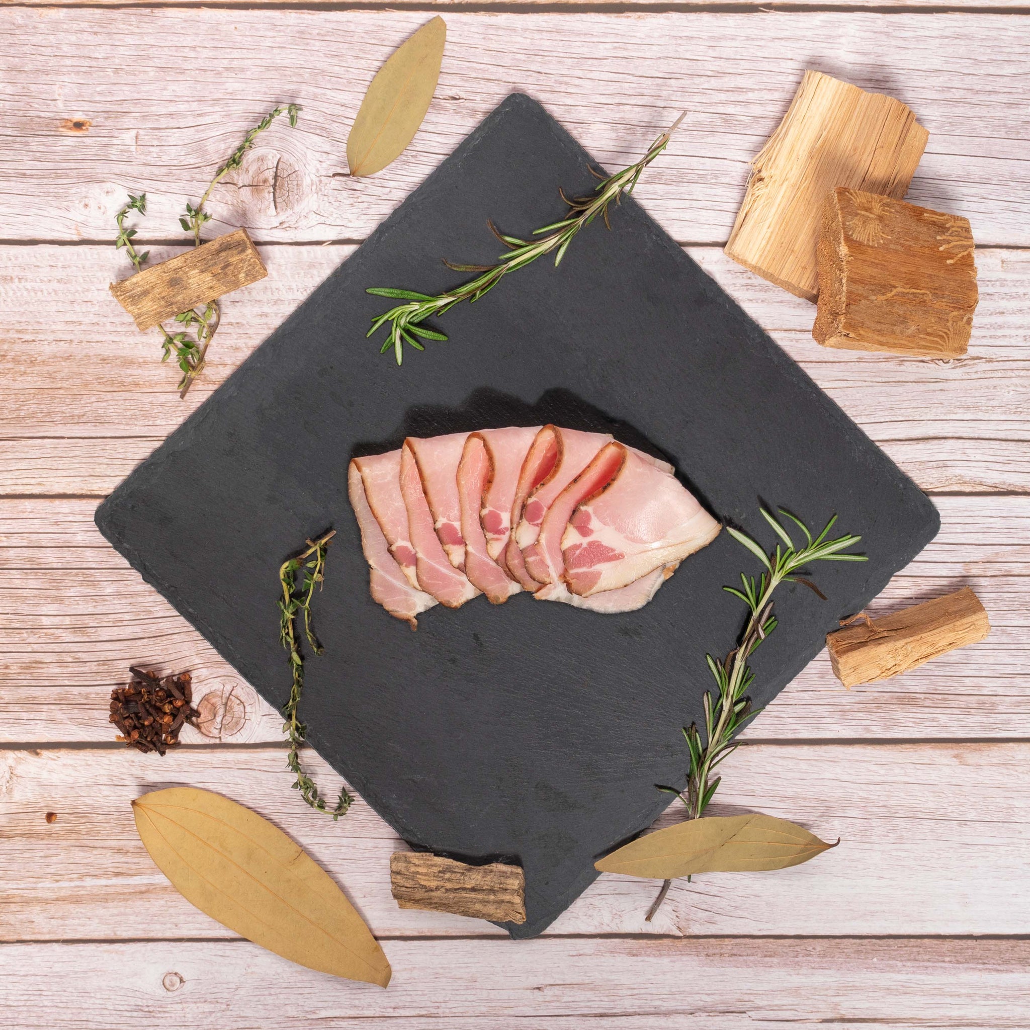 Slices of Specialty Smoked Ham on Stone Plate with Bay Leaves, Rosemary, Thyme, Cloves, Applewood chips decorating the side, against a marble background