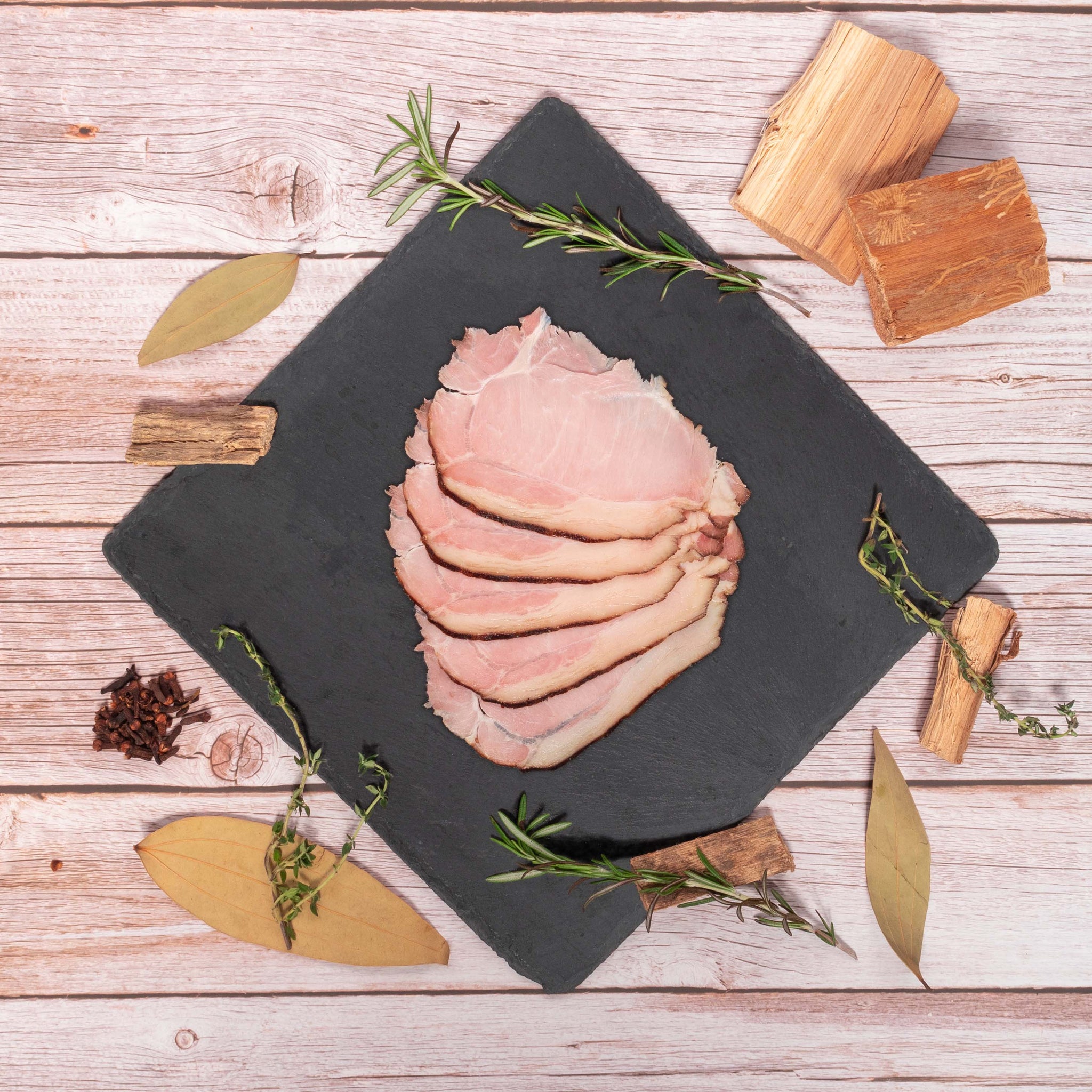 Chestnut-fed Smoked Ham on Stone Plate with Bay Leaves, Rosemary, Thyme, Cloves, Applewood chips decorating the side.