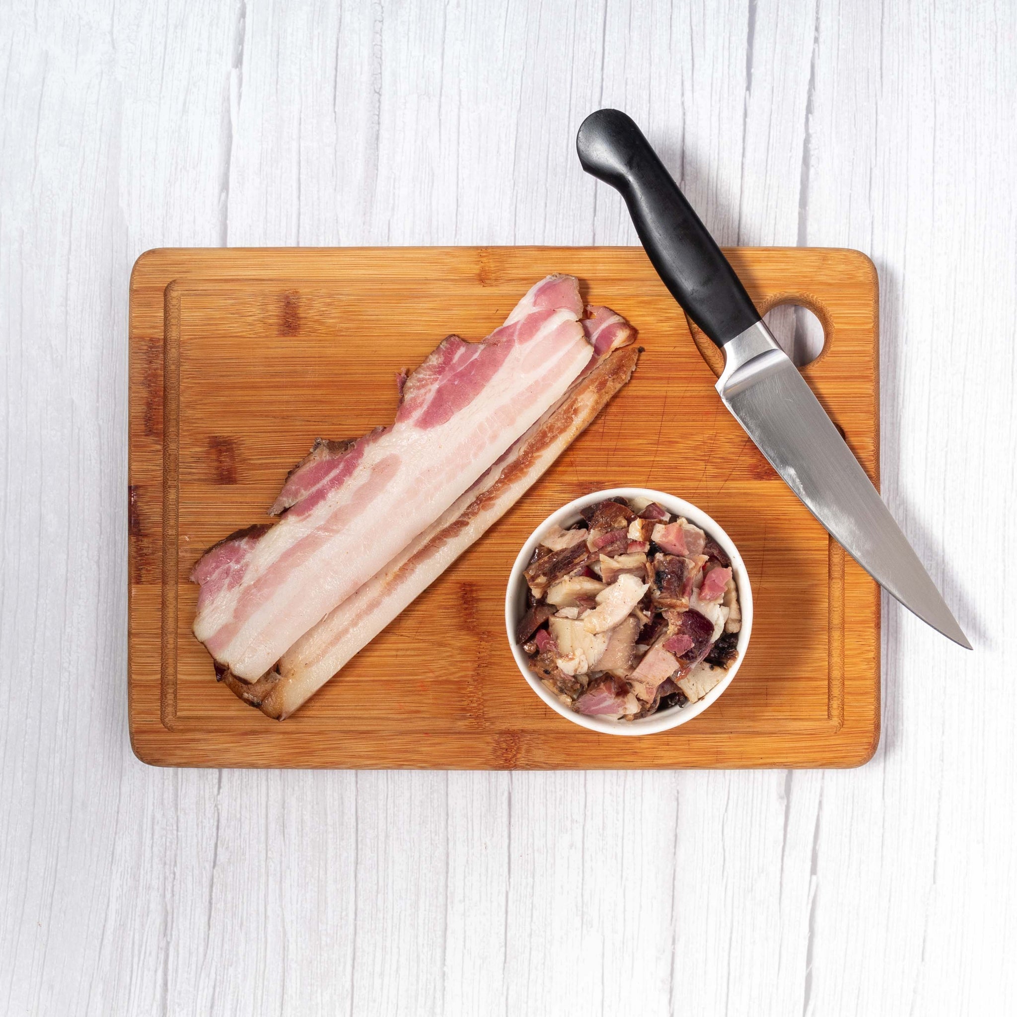 Slices of Smoked Bacon with Chopped up Nitrite-free Smoked Bacon Bits on the side, with a silver knife next to it, all placed on top a wooden chopping board.